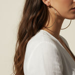 This pair of large gold hoop earrings features a smooth, uninterrupted circular silhouette crafted from a sleek round wire, offering a timeless, versatile shape that works for both everyday wear and dressed-up occasions. The hoops close securely with a hinged snap-back mechanism, ensuring a comfortable, secure fit at the earlobe. Photographed from multiple angles against a clean, neutral background, the dramatic diagonal shadow and bright directional lighting highlight the hoops' polished, reflective gold f