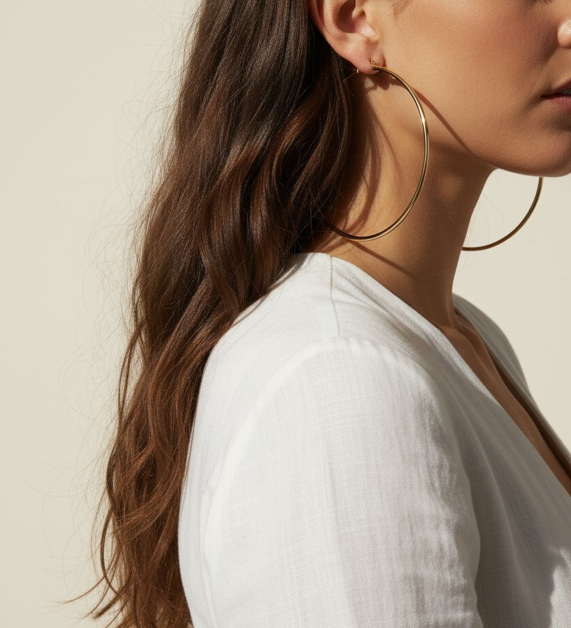 This pair of large gold hoop earrings features a smooth, uninterrupted circular silhouette crafted from a sleek round wire, offering a timeless, versatile shape that works for both everyday wear and dressed-up occasions. The hoops close securely with a hinged snap-back mechanism, ensuring a comfortable, secure fit at the earlobe. Photographed from multiple angles against a clean, neutral background, the dramatic diagonal shadow and bright directional lighting highlight the hoops' polished, reflective gold f