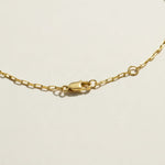 This gold-tone paperclip chain necklace is centered on a square-cut pendant with a large clear crystal set in a polished bezel. A pavé halo of tiny clear stones surrounds the center stone and adds sparkle at every angle. The first frame is a straight-on flat lay on a warm cream surface, showing the full pendant face and the pavé border. The second frame is an angled close-up of the pendant’s slotted, gridded side walls and the pavé setting. The third frame is a detail of the clasp, a lobster clasp joined to