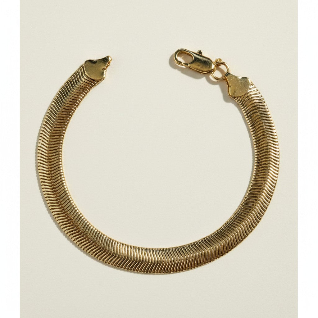 This gold bracelet features a wide, flat herringbone chain, prized for its smooth, snake-like texture and high-shine reflective finish. The tightly woven flat links lie sleek against the wrist, catching the light with every movement for a bold, glossy effect. It fastens securely with a lobster clasp closure, giving the piece a polished, finished look ideal for stacking or wearing solo. Photographed flat against a clean, neutral background, the bracelet is shown in a soft circular drape that highlights the c