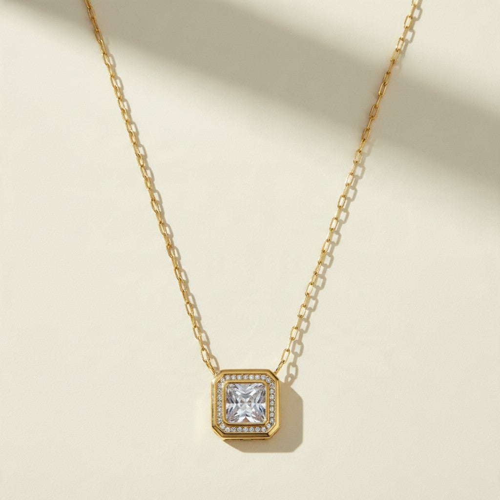 This gold-tone paperclip chain necklace is centered on a square-cut pendant with a large clear crystal set in a polished bezel. A pavé halo of tiny clear stones surrounds the center stone and adds sparkle at every angle. The first frame is a straight-on flat lay on a warm cream surface, showing the full pendant face and the pavé border. The second frame is an angled close-up of the pendant’s slotted, gridded side walls and the pavé setting. The third frame is a detail of the clasp, a lobster clasp joined to