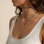 This series of portraits shows a gold paperclip link necklace worn at the collarbone, featuring an elongated oval-link chain that centers on a polished circular ring clasp as a modern, sculptural focal point. Captured from front-facing and side-profile angles against a crisp white tank top, the necklace is shown at true scale, giving shoppers a clear sense of its bold link proportions and how the round clasp element sits as an intentional design detail rather than a hidden closure