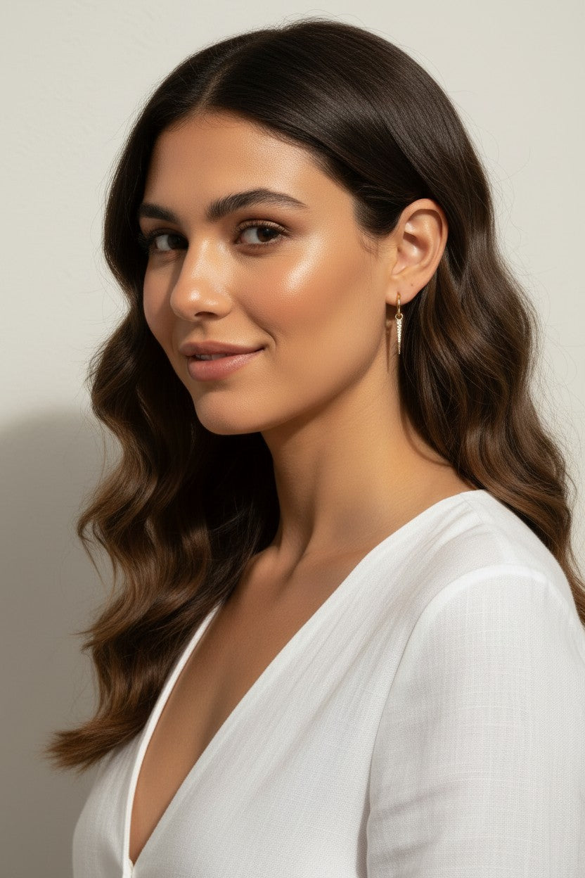 This series of close-up portraits shows the CZ spear charm attached to a small gold seamless hoop, worn at the lobe for a customized, layered earring look. Captured from multiple angles against soft waved hair and a crisp white top, the charm's graduated cubic zirconia setting and tapered point are shown at true scale, giving shoppers a clear sense of drop length and how the 18k gold overlay charm pairs with the 9mm or 11mm hoop.