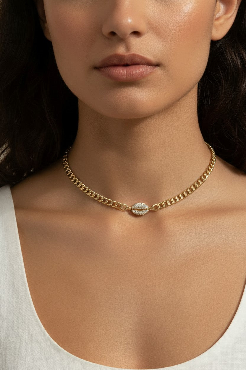 This on-model close-up showcases a gold curb chain choker sitting neatly at the base of the neck, with a pavé crystal cowrie shell charm resting just above the collarbone as the eye-catching centerpiece. Styled with a white scoop-neck top, the choker's short length and close fit highlight how the chunky curb links and sparkling shell charm create a polished, versatile accent — equally suited to everyday wear or elevated occasions. The soft, neutral-toned backdrop and natural lighting emphasize the necklace'