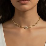 This on-model close-up showcases a gold curb chain choker sitting neatly at the base of the neck, with a pavé crystal cowrie shell charm resting just above the collarbone as the eye-catching centerpiece. Styled with a white scoop-neck top, the choker's short length and close fit highlight how the chunky curb links and sparkling shell charm create a polished, versatile accent — equally suited to everyday wear or elevated occasions. The soft, neutral-toned backdrop and natural lighting emphasize the necklace'