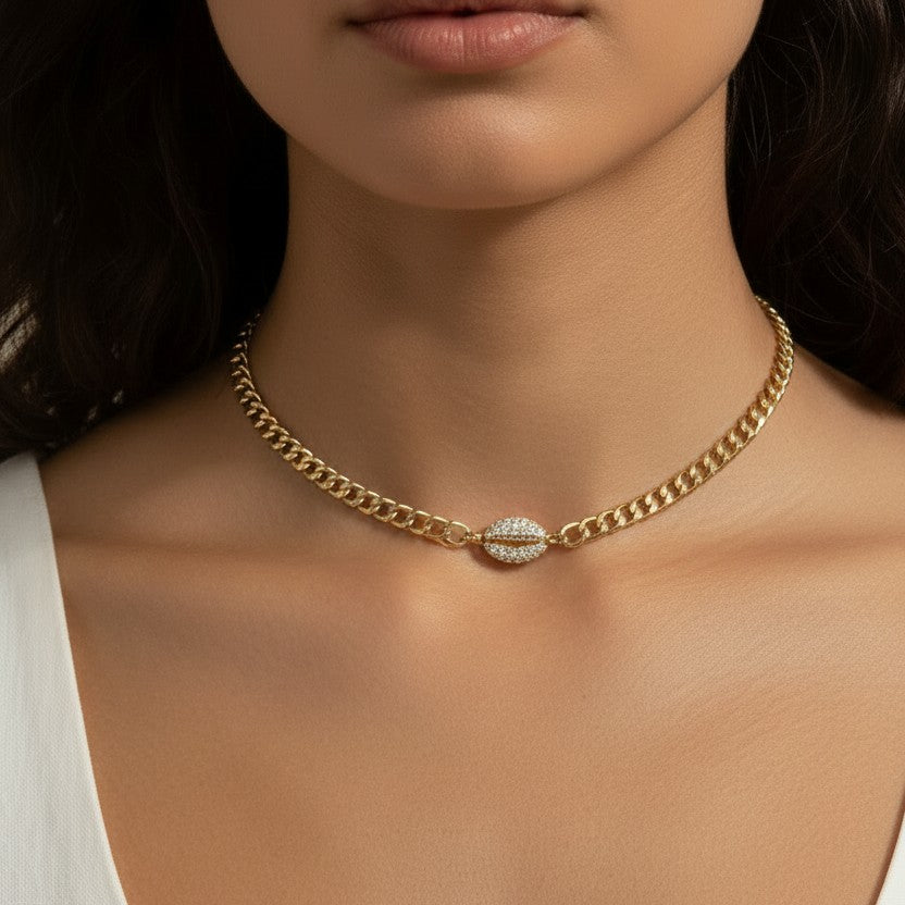 This on-model close-up showcases a gold curb chain choker sitting neatly at the base of the neck, with a pavé crystal cowrie shell charm resting just above the collarbone as the eye-catching centerpiece. Styled with a white scoop-neck top, the choker's short length and close fit highlight how the chunky curb links and sparkling shell charm create a polished, versatile accent — equally suited to everyday wear or elevated occasions. The soft, neutral-toned backdrop and natural lighting emphasize the necklace'