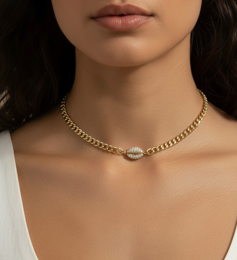 This on-model close-up showcases a gold curb chain choker sitting neatly at the base of the neck, with a pavé crystal cowrie shell charm resting just above the collarbone as the eye-catching centerpiece. Styled with a white scoop-neck top, the choker's short length and close fit highlight how the chunky curb links and sparkling shell charm create a polished, versatile accent — equally suited to everyday wear or elevated occasions. The soft, neutral-toned backdrop and natural lighting emphasize the necklace'