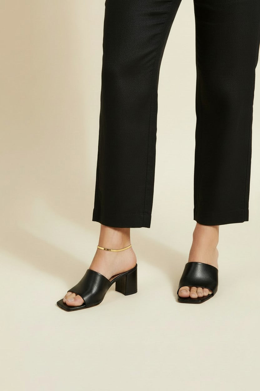 This on-model lifestyle photo showcases a delicate gold anklet worn at the ankle bone, paired with black square-toe block heel mule sandals and tailored black cropped trousers. The slim gold chain sits neatly against bare skin, offering a subtle, elegant accent that bridges the gap between hemline and shoe. Shot from a side angle against a warm neutral backdrop, the styling highlights how the anklet layers effortlessly into a polished, minimalist office-to-evening look. The soft, warm-toned background and n