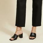 This on-model lifestyle photo showcases a delicate gold anklet worn at the ankle bone, paired with black square-toe block heel mule sandals and tailored black cropped trousers. The slim gold chain sits neatly against bare skin, offering a subtle, elegant accent that bridges the gap between hemline and shoe. Shot from a side angle against a warm neutral backdrop, the styling highlights how the anklet layers effortlessly into a polished, minimalist office-to-evening look. The soft, warm-toned background and n