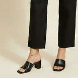 This on-model lifestyle photo showcases a delicate gold anklet worn at the ankle bone, paired with black square-toe block heel mule sandals and tailored black cropped trousers. The slim gold chain sits neatly against bare skin, offering a subtle, elegant accent that bridges the gap between hemline and shoe. Shot from a side angle against a warm neutral backdrop, the styling highlights how the anklet layers effortlessly into a polished, minimalist office-to-evening look. The soft, warm-toned background and n