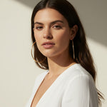 This series of lifestyle portraits shows large gold hoop earrings worn at the ear, their smooth round-wire silhouette shown at true scale against soft waved hair and warm, sunlit skin. Styled with a plunging white wrap top in both an indoor studio setting and an outdoor balcony scene, the hoops are captured from front-facing, side-profile, and close-up angles, giving shoppers a comprehensive view of drop length, diameter, and fit. 