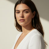 This series of lifestyle portraits shows large gold hoop earrings worn at the ear, their smooth round-wire silhouette shown at true scale against soft waved hair and warm, sunlit skin. Styled with a plunging white wrap top in both an indoor studio setting and an outdoor balcony scene, the hoops are captured from front-facing, side-profile, and close-up angles, giving shoppers a comprehensive view of drop length, diameter, and fit. 