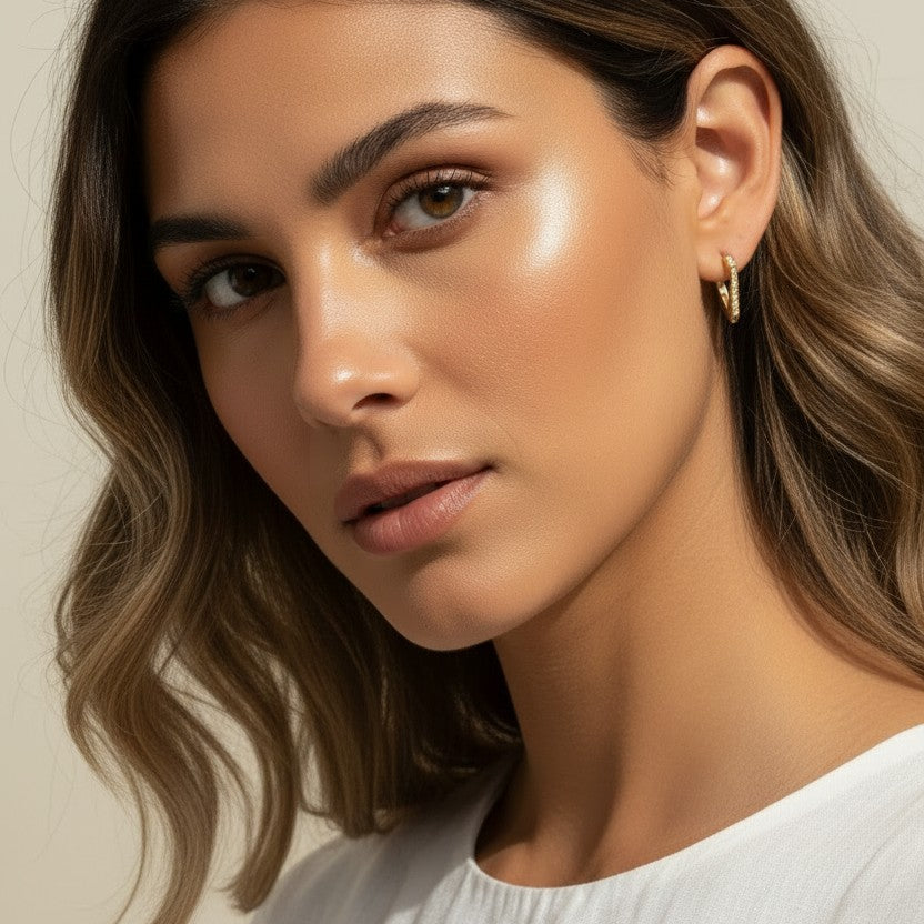A close-up portrait of a woman with wavy brown hair wearing a delicate gold hoop earring, set against a soft beige background.