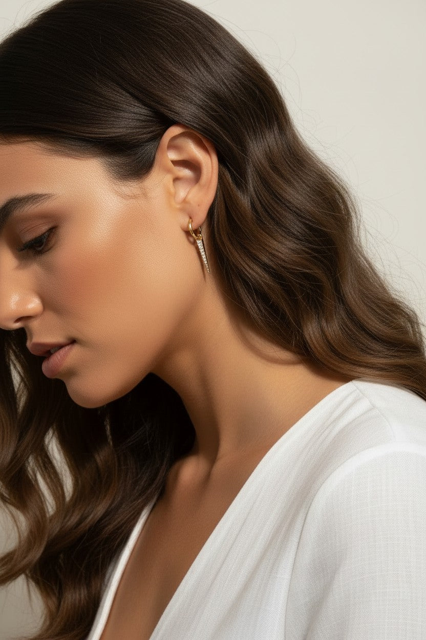 This series of close-up portraits shows the CZ spear charm attached to a small gold seamless hoop, worn at the lobe for a customized, layered earring look. Captured from multiple angles against soft waved hair and a crisp white top, the charm's graduated cubic zirconia setting and tapered point are shown at true scale, giving shoppers a clear sense of drop length and how the 18k gold overlay charm pairs with the 9mm or 11mm hoop.