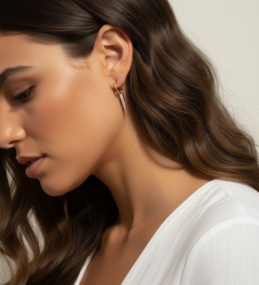 This series of close-up portraits shows the CZ spear charm attached to a small gold seamless hoop, worn at the lobe for a customized, layered earring look. Captured from multiple angles against soft waved hair and a crisp white top, the charm's graduated cubic zirconia setting and tapered point are shown at true scale, giving shoppers a clear sense of drop length and how the 18k gold overlay charm pairs with the 9mm or 11mm hoop.