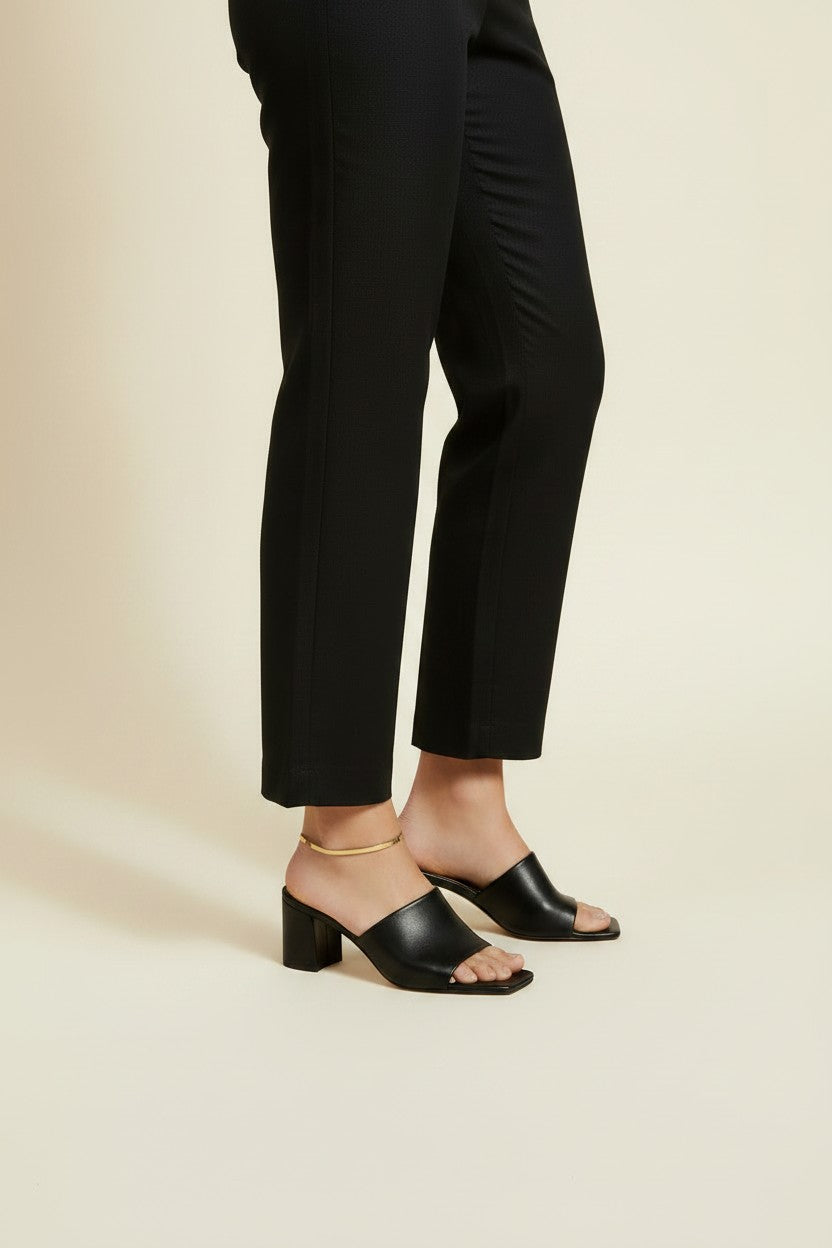 This on-model lifestyle photo showcases a delicate gold anklet worn at the ankle bone, paired with black square-toe block heel mule sandals and tailored black cropped trousers. The slim gold chain sits neatly against bare skin, offering a subtle, elegant accent that bridges the gap between hemline and shoe. Shot from a side angle against a warm neutral backdrop, the styling highlights how the anklet layers effortlessly into a polished, minimalist office-to-evening look. The soft, warm-toned background and n