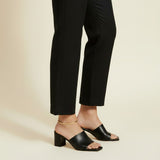 This on-model lifestyle photo showcases a delicate gold anklet worn at the ankle bone, paired with black square-toe block heel mule sandals and tailored black cropped trousers. The slim gold chain sits neatly against bare skin, offering a subtle, elegant accent that bridges the gap between hemline and shoe. Shot from a side angle against a warm neutral backdrop, the styling highlights how the anklet layers effortlessly into a polished, minimalist office-to-evening look. The soft, warm-toned background and n