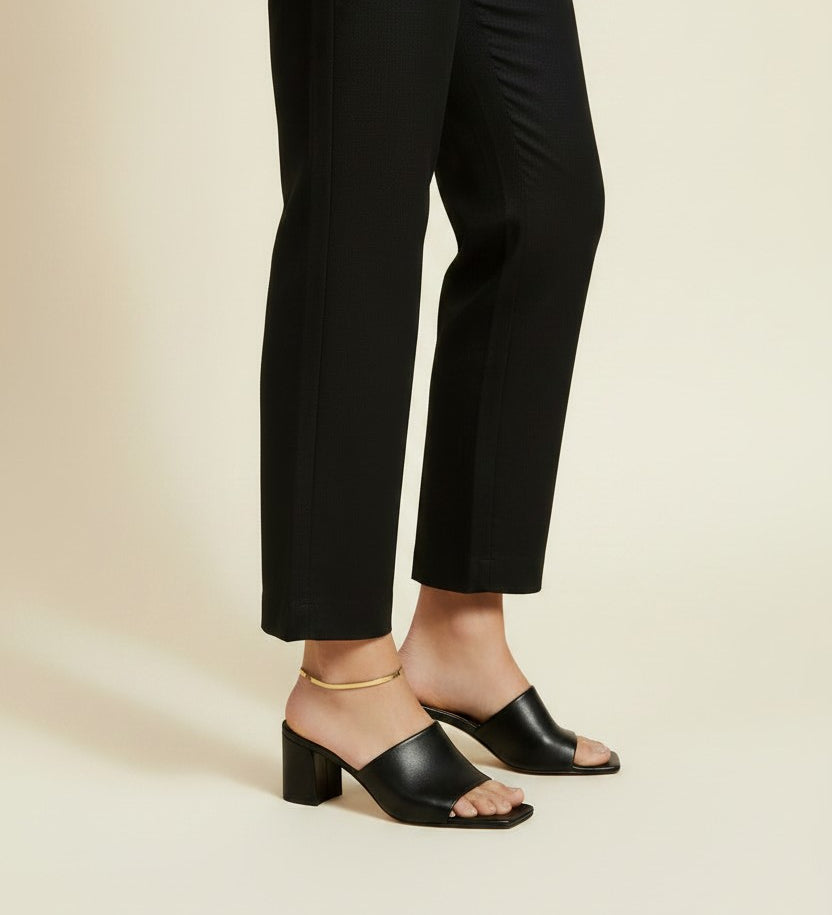 This on-model lifestyle photo showcases a delicate gold anklet worn at the ankle bone, paired with black square-toe block heel mule sandals and tailored black cropped trousers. The slim gold chain sits neatly against bare skin, offering a subtle, elegant accent that bridges the gap between hemline and shoe. Shot from a side angle against a warm neutral backdrop, the styling highlights how the anklet layers effortlessly into a polished, minimalist office-to-evening look. The soft, warm-toned background and n