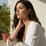 This series of lifestyle portraits shows large gold hoop earrings worn at the ear, their smooth round-wire silhouette shown at true scale against soft waved hair and warm, sunlit skin. Styled with a plunging white wrap top in both an indoor studio setting and an outdoor balcony scene, the hoops are captured from front-facing, side-profile, and close-up angles, giving shoppers a comprehensive view of drop length, diameter, and fit. Bold natural sunlight and dramatic shadow accents create a warm, sun-drenched