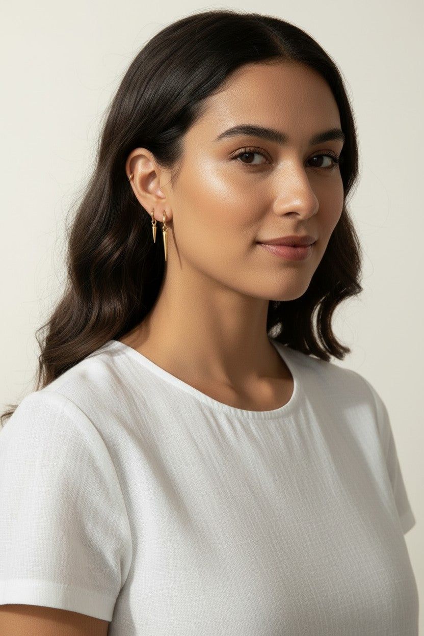 Gold Spike Charm Hoop Earrings Worn On-Model, dangling triangular spike charm at the bottom, worn together with a thin ear cuff along the upper ear for a curated, layered ear stack look. Captured from front-facing, side-profile, and close-up angles, the earrings are shown at true scale against soft waved hair and a crisp white top, giving shoppers a clear sense of the charm's drop length and how the pieces sit together