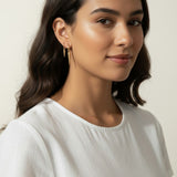 Gold Spike Charm Hoop Earrings Worn On-Model, dangling triangular spike charm at the bottom, worn together with a thin ear cuff along the upper ear for a curated, layered ear stack look. Captured from front-facing, side-profile, and close-up angles, the earrings are shown at true scale against soft waved hair and a crisp white top, giving shoppers a clear sense of the charm's drop length and how the pieces sit together