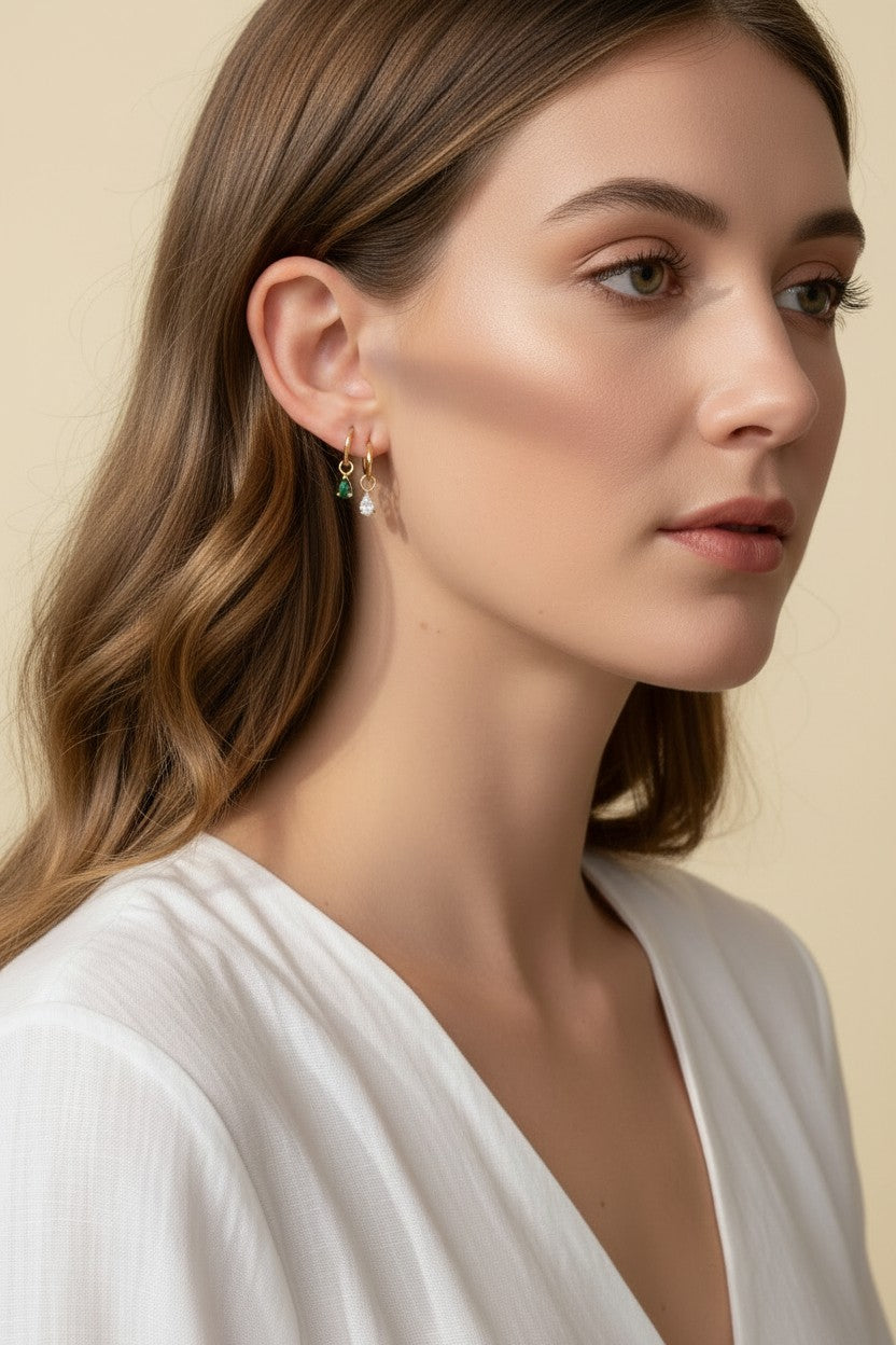 This close-up profile shot shows a pair of small gold hoop earrings, each accented with a dangling pear-cut crystal charm — one in emerald green, one in clear crystal — worn together along the lower earlobe. The delicate hoops sit close to the ear with the faceted charms swinging just below, shown at true scale against soft waved hair and a white wrap top for a clear sense of drop length and fit. Warm, soft lighting creates a clean, romantic mood, highlighting how the mixed emerald-and-clear pairing offers 