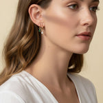 This close-up profile shot shows a pair of small gold hoop earrings, each accented with a dangling pear-cut crystal charm — one in emerald green, one in clear crystal — worn together along the lower earlobe. The delicate hoops sit close to the ear with the faceted charms swinging just below, shown at true scale against soft waved hair and a white wrap top for a clear sense of drop length and fit. Warm, soft lighting creates a clean, romantic mood, highlighting how the mixed emerald-and-clear pairing offers 
