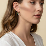 This close-up profile shot shows a pair of small gold hoop earrings, each accented with a dangling pear-cut crystal charm — one in emerald green, one in clear crystal — worn together along the lower earlobe. The delicate hoops sit close to the ear with the faceted charms swinging just below, shown at true scale against soft waved hair and a white wrap top for a clear sense of drop length and fit. Warm, soft lighting creates a clean, romantic mood, highlighting how the mixed emerald-and-clear pairing offers 