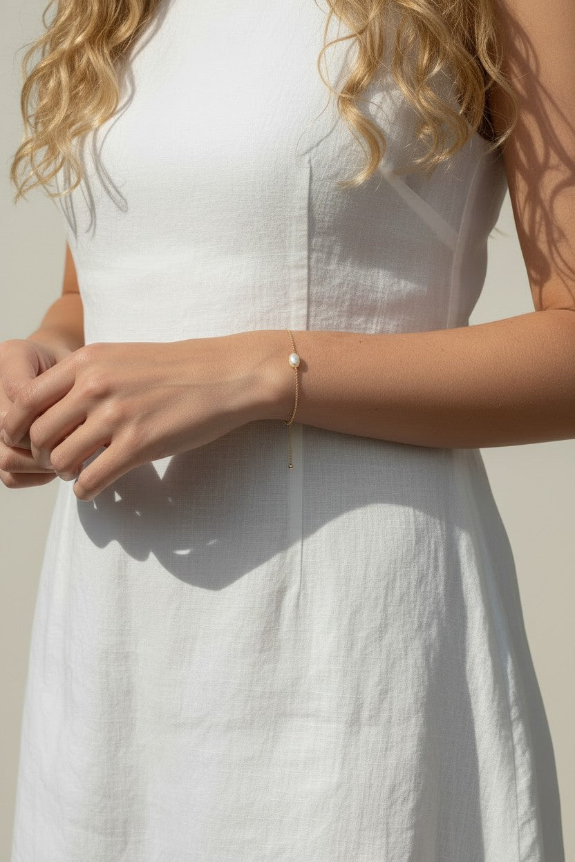 This series of lifestyle portraits shows a delicate gold chain bracelet resting on the wrist, featuring a single freshwater pearl set off-center along a fine cable chain. Styled with a flowing white linen dress in warm, sunlit settings, the bracelet is shown at true scale from multiple angles, highlighting its dainty adjustable drop chain and the soft, organic luster of the pearl against sun-kissed skin. 