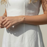 This series of lifestyle portraits shows a delicate gold chain bracelet resting on the wrist, featuring a single freshwater pearl set off-center along a fine cable chain. Styled with a flowing white linen dress in warm, sunlit settings, the bracelet is shown at true scale from multiple angles, highlighting its dainty adjustable drop chain and the soft, organic luster of the pearl against sun-kissed skin. 