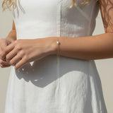 This series of lifestyle portraits shows a delicate gold chain bracelet resting on the wrist, featuring a single freshwater pearl set off-center along a fine cable chain. Styled with a flowing white linen dress in warm, sunlit settings, the bracelet is shown at true scale from multiple angles, highlighting its dainty adjustable drop chain and the soft, organic luster of the pearl against sun-kissed skin. 