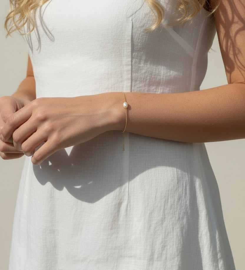 This series of lifestyle portraits shows a delicate gold chain bracelet resting on the wrist, featuring a single freshwater pearl set off-center along a fine cable chain. Styled with a flowing white linen dress in warm, sunlit settings, the bracelet is shown at true scale from multiple angles, highlighting its dainty adjustable drop chain and the soft, organic luster of the pearl against sun-kissed skin. 