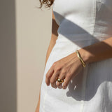 This sunlit close-up shows a gold herringbone chain bracelet wrapped around the wrist, its smooth, flat links catching warm natural light for a rich, reflective shine. Styled with a crisp white linen dress and a chunky gold dome ring on the finger, the bracelet is shown at true scale, giving shoppers a clear sense of its fit and how it drapes against the skin. Bold, graphic shadows and golden-hour lighting create a striking editorial mood, while the relaxed hand pose highlights how the sleek herringbone tex