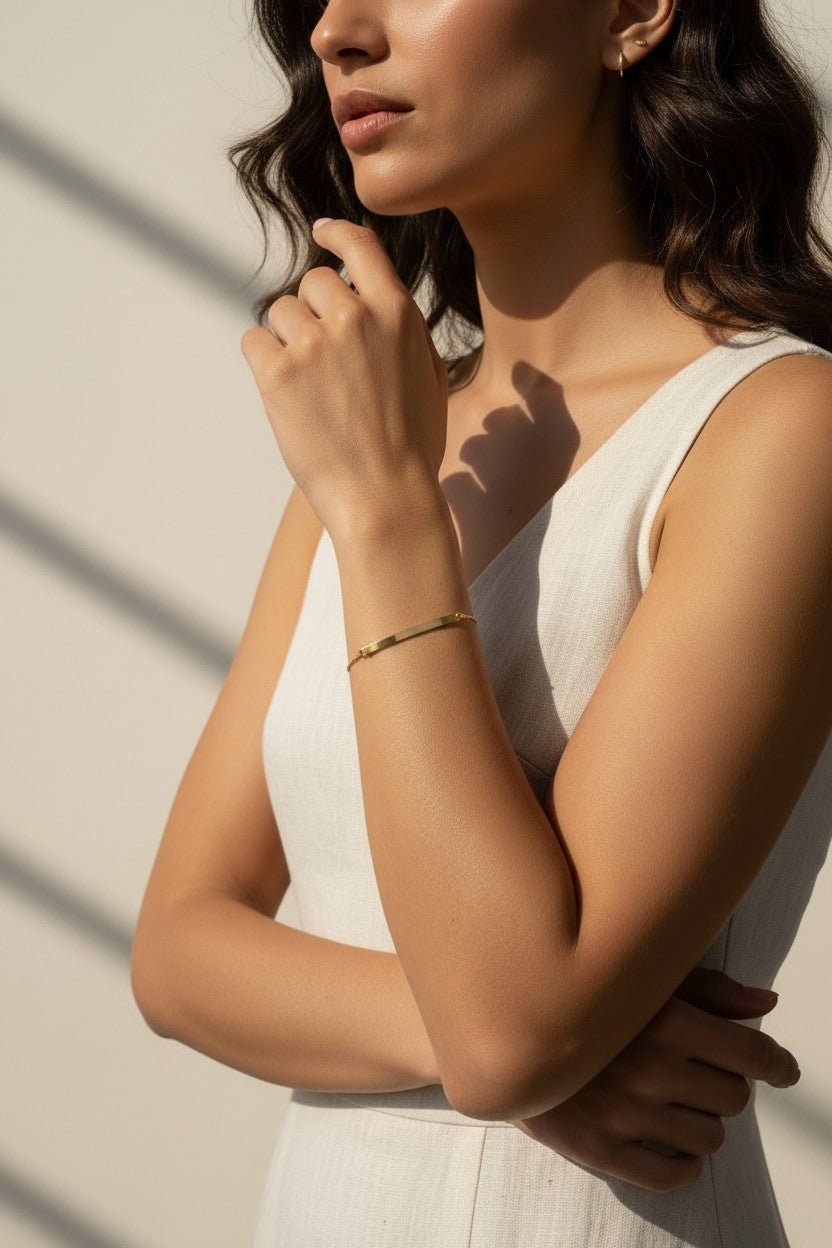 This editorial close-up captures a slim gold bracelet resting on the wrist, featuring a slender bangle band accented with a raised horizontal bar detail. The model's crossed-arm pose highlights the bracelet's sleek, low-profile fit against the skin, styled with a draped white dress and small gold hoop earrings for an effortlessly polished look. Warm, directional sunlight and bold graphic shadow lines across the frame create a striking editorial mood, while emphasizing the bracelet's smooth, reflective 