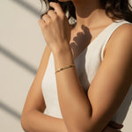 This editorial close-up captures a slim gold bracelet resting on the wrist, featuring a slender bangle band accented with a raised horizontal bar detail. The model's crossed-arm pose highlights the bracelet's sleek, low-profile fit against the skin, styled with a draped white dress and small gold hoop earrings for an effortlessly polished look. Warm, directional sunlight and bold graphic shadow lines across the frame create a striking editorial mood, while emphasizing the bracelet's smooth, reflective 