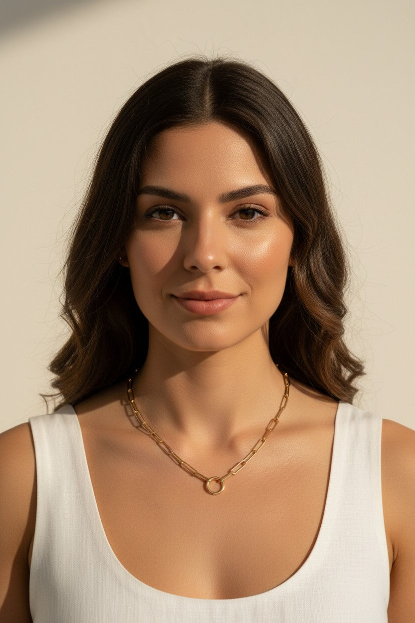 This series of portraits shows a gold paperclip link necklace worn at the collarbone, featuring an elongated oval-link chain that centers on a polished circular ring clasp as a modern, sculptural focal point. Captured from front-facing and side-profile angles against a crisp white tank top, the necklace is shown at true scale, giving shoppers a clear sense of its bold link proportions and how the round clasp element sits as an intentional design detail rather than a hidden closure