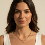 This series of portraits shows a gold paperclip link necklace worn at the collarbone, featuring an elongated oval-link chain that centers on a polished circular ring clasp as a modern, sculptural focal point. Captured from front-facing and side-profile angles against a crisp white tank top, the necklace is shown at true scale, giving shoppers a clear sense of its bold link proportions and how the round clasp element sits as an intentional design detail rather than a hidden closure