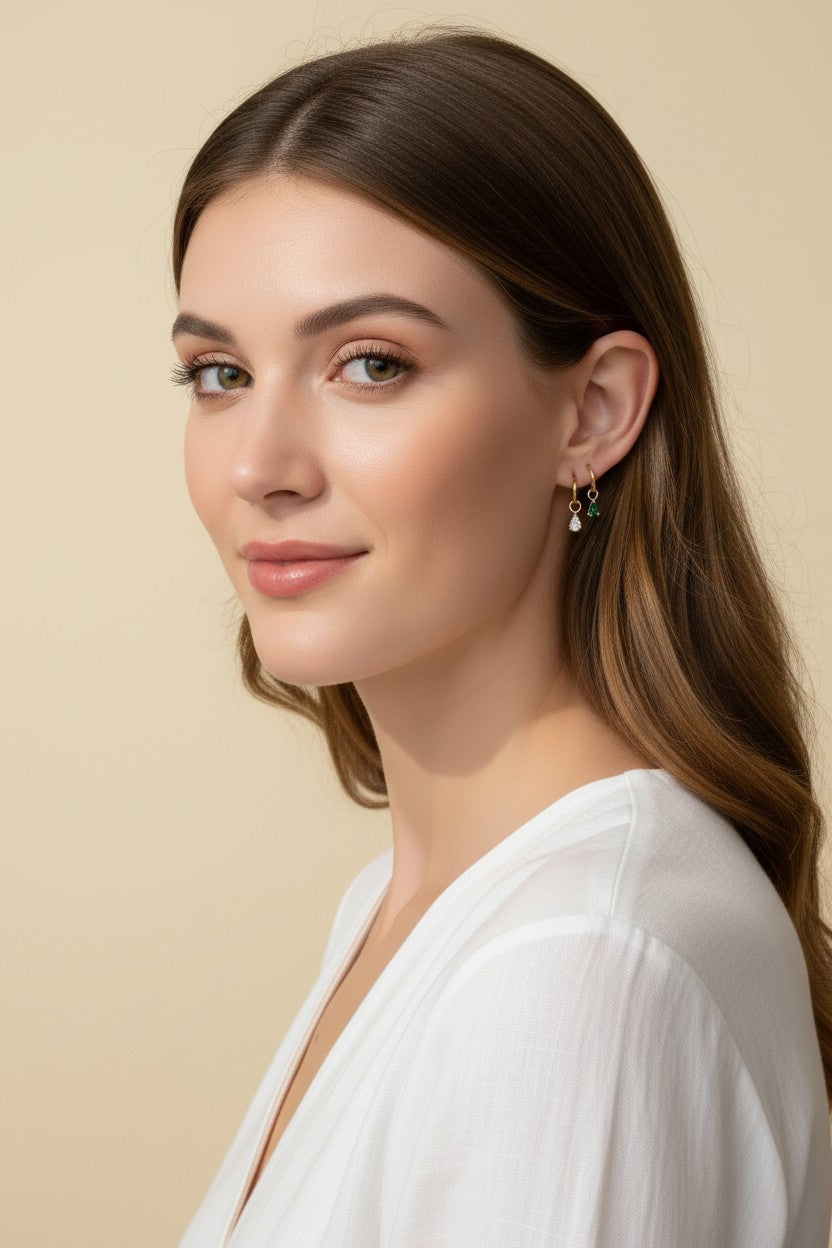 This close-up profile shot shows a pair of small gold hoop earrings, each accented with a dangling pear-cut crystal charm — one in emerald green, one in clear crystal — worn together along the lower earlobe. The delicate hoops sit close to the ear with the faceted charms swinging just below, shown at true scale against soft waved hair and a white wrap top for a clear sense of drop length and fit. Warm, soft lighting creates a clean, romantic mood, highlighting how the mixed emerald-and-clear pairing offers 