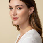 This close-up profile shot shows a pair of small gold hoop earrings, each accented with a dangling pear-cut crystal charm — one in emerald green, one in clear crystal — worn together along the lower earlobe. The delicate hoops sit close to the ear with the faceted charms swinging just below, shown at true scale against soft waved hair and a white wrap top for a clear sense of drop length and fit. Warm, soft lighting creates a clean, romantic mood, highlighting how the mixed emerald-and-clear pairing offers 