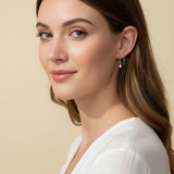 This close-up profile shot shows a pair of small gold hoop earrings, each accented with a dangling pear-cut crystal charm — one in emerald green, one in clear crystal — worn together along the lower earlobe. The delicate hoops sit close to the ear with the faceted charms swinging just below, shown at true scale against soft waved hair and a white wrap top for a clear sense of drop length and fit. Warm, soft lighting creates a clean, romantic mood, highlighting how the mixed emerald-and-clear pairing offers 