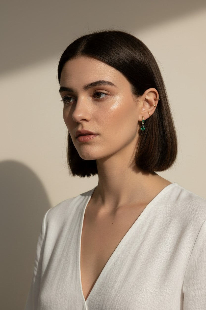 This close-up profile shot shows the emerald green marquise drop earring worn on the lobe, its faceted stone catching soft light for a rich, jewel-toned sparkle. Styled with a sleek center-part bob and layered with multiple thin gold ear cuffs along the upper ear, the earring is shown at true scale, giving shoppers a clear sense of its drop length and how it moves against the jawline. 