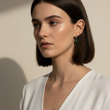 This close-up profile shot shows the emerald green marquise drop earring worn on the lobe, its faceted stone catching soft light for a rich, jewel-toned sparkle. Styled with a sleek center-part bob and layered with multiple thin gold ear cuffs along the upper ear, the earring is shown at true scale, giving shoppers a clear sense of its drop length and how it moves against the jawline. 