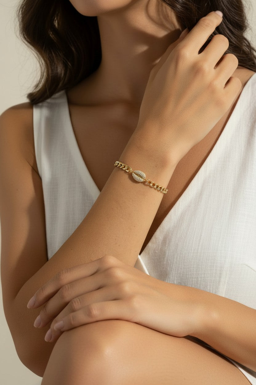 This close-up lifestyle shot shows a gold curb chain bracelet resting elegantly on the wrist, with the pavé crystal cowrie shell charm sitting front and center as the piece's focal point. Styled with a white linen wrap top, the bracelet's bold chain links and sparkling shell charm are shown at true scale against warm skin tones, giving shoppers a clear sense of fit and drape. Soft, warm natural lighting and a relaxed hand-to-collarbone pose lend the image an intimate, editorial feel, highlighting the interp