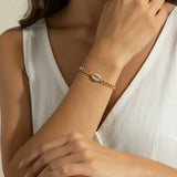 This close-up lifestyle shot shows a gold curb chain bracelet resting elegantly on the wrist, with the pavé crystal cowrie shell charm sitting front and center as the piece's focal point. Styled with a white linen wrap top, the bracelet's bold chain links and sparkling shell charm are shown at true scale against warm skin tones, giving shoppers a clear sense of fit and drape. Soft, warm natural lighting and a relaxed hand-to-collarbone pose lend the image an intimate, editorial feel, highlighting the interp
