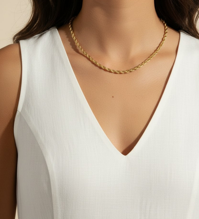 This close-up profile shot shows a gold rope chain necklace resting neatly at collarbone length, its tightly twisted, braided texture catching soft studio light for a warm, polished shine. Styled with a sleeveless white V-neck top, the necklace is shown at true scale against the neckline, giving shoppers a clear sense of its length and how it sits on the body — versatile enough for both everyday wear and elevated occasions. The clean, neutral background and gentle side lighting keep the focus on the chain's