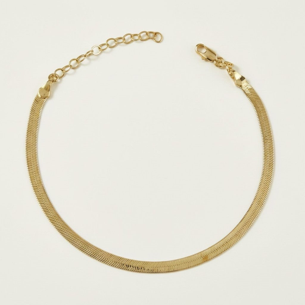 This delicate gold anklet features a sleek herringbone chain design, prized for its flat, snake-like texture and high-shine reflective finish. The tightly woven flat links lie smooth and comfortable against the skin, catching the light with every movement. It fastens securely with a classic lobster clasp closure and includes a 2-inch extender chain with a small round jump ring, allowing for an adjustable, customizable fit around the ankle. Photographed against a clean, neutral off-white background, the piec