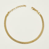 This delicate gold anklet features a sleek herringbone chain design, prized for its flat, snake-like texture and high-shine reflective finish. The tightly woven flat links lie smooth and comfortable against the skin, catching the light with every movement. It fastens securely with a classic lobster clasp closure and includes a 2-inch extender chain with a small round jump ring, allowing for an adjustable, customizable fit around the ankle. Photographed against a clean, neutral off-white background, the piec