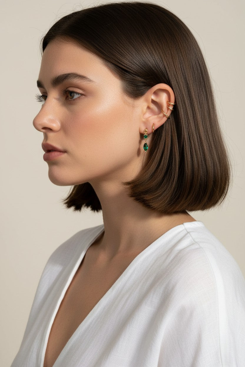 This close-up profile shot shows the emerald green marquise drop earring worn on the lobe, its faceted stone catching soft light for a rich, jewel-toned sparkle. Styled with a sleek center-part bob and layered with multiple thin gold ear cuffs along the upper ear, the earring is shown at true scale, giving shoppers a clear sense of its drop length and how it moves against the jawline. The warm, dramatic side lighting and minimalist white top create a polished, editorial mood, highlighting how the emerald dr