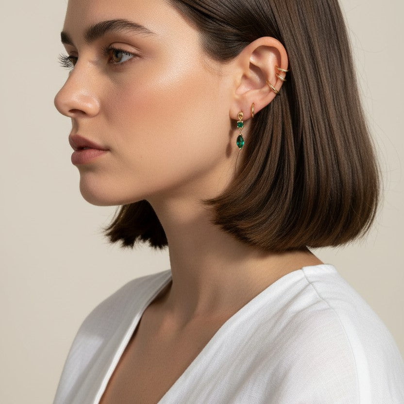 This close-up profile shot shows the emerald green marquise drop earring worn on the lobe, its faceted stone catching soft light for a rich, jewel-toned sparkle. Styled with a sleek center-part bob and layered with multiple thin gold ear cuffs along the upper ear, the earring is shown at true scale, giving shoppers a clear sense of its drop length and how it moves against the jawline. The warm, dramatic side lighting and minimalist white top create a polished, editorial mood, highlighting how the emerald dr
