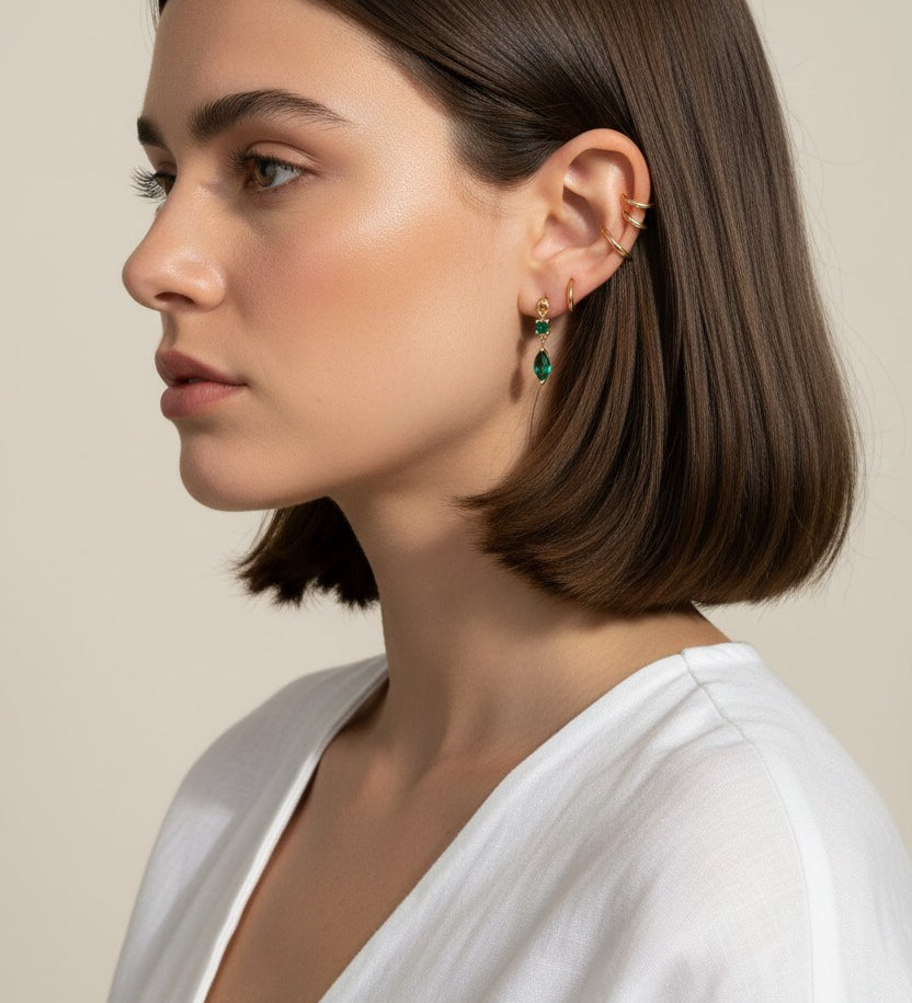 This close-up profile shot shows the emerald green marquise drop earring worn on the lobe, its faceted stone catching soft light for a rich, jewel-toned sparkle. Styled with a sleek center-part bob and layered with multiple thin gold ear cuffs along the upper ear, the earring is shown at true scale, giving shoppers a clear sense of its drop length and how it moves against the jawline. The warm, dramatic side lighting and minimalist white top create a polished, editorial mood, highlighting how the emerald dr